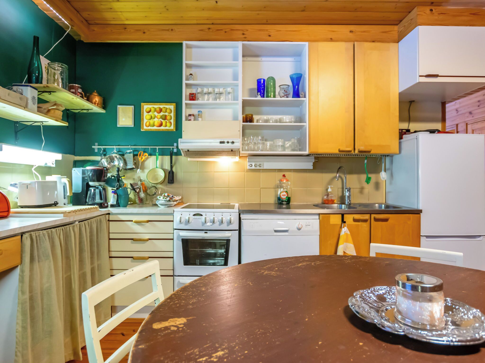 Een keuken met witte kasten, een fornuis, een vaatwasser en een koelkast. Er staat een eettafel met een kaars op een dienblad.