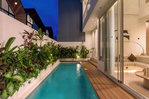 A modern outdoor pool area with a wooden deck, surrounded by lush greenery and a glass wall leading to a living area with a sofa and coffee table. - Villa Fresko 2 by Alfred in Bali (Munggu)