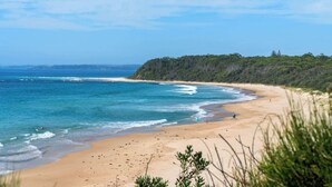Uma praia com águas azuis cristalinas, areal arenoso e um penhasco coberto de vegetação.