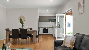 Eine moderne Küche mit weißen Schränken, ein Essbereich mit Holztisch und -stühlen sowie eine Ledersofa mit Sitzkissen.