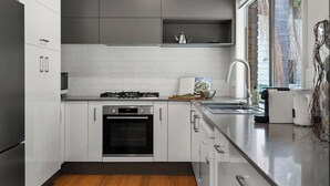 Eine moderne Küche mit weißen Schränken, einem Edelstahlkühlschrank, einem eingebauten Backofen, einem Gasherd und einem Fenster mit Blick auf Grünflächen.