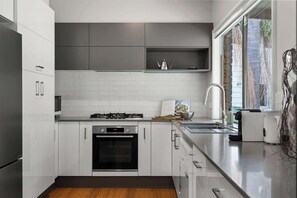 Eine moderne Küche mit weißen Schränken, einem Edelstahlkühlschrank, einem eingebauten Backofen, einem Gasherd und einem Fenster mit Blick auf Grünflächen.