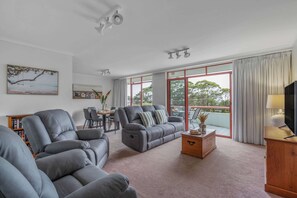 Ein Wohnzimmer mit grauen Sofas, einem Holztisch, einem Fernseher und einem Balkon mit Vorhängen.