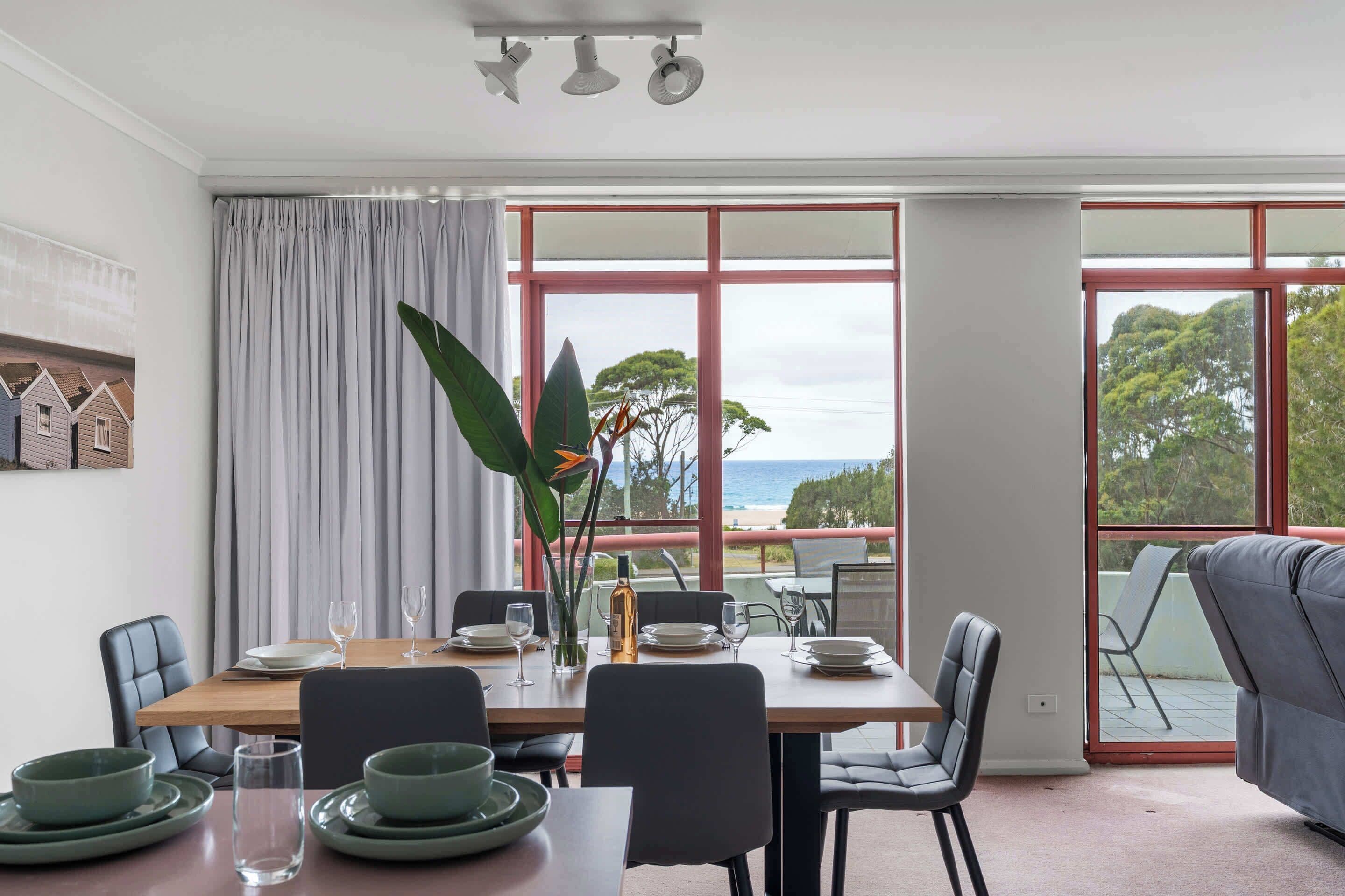 A dining area with a table set for four, large windows with a view, and a grey sofa in the foreground.