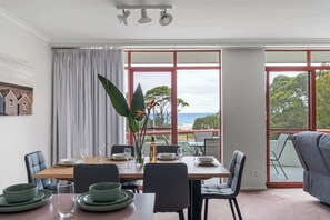 Ein Essbereich mit Tisch für vier Personen, großen Fenstern mit Blick und einem grauen Sofa im Vordergrund.