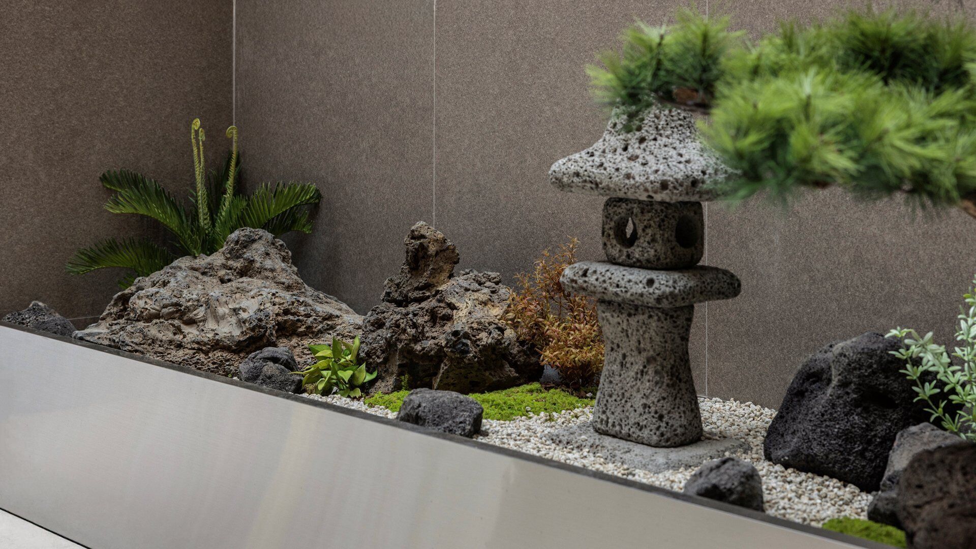Een rotstuin met een stenen sculptuur, diverse planten en een muur op de achtergrond.