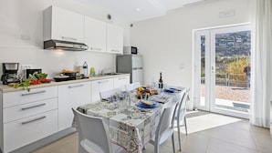 A modern kitchen with white cabinets, a dining table set with plates, glasses, and a bottle, and a view of a hillside through a glass door. - Villa al Fiume (Nago-Torbole)