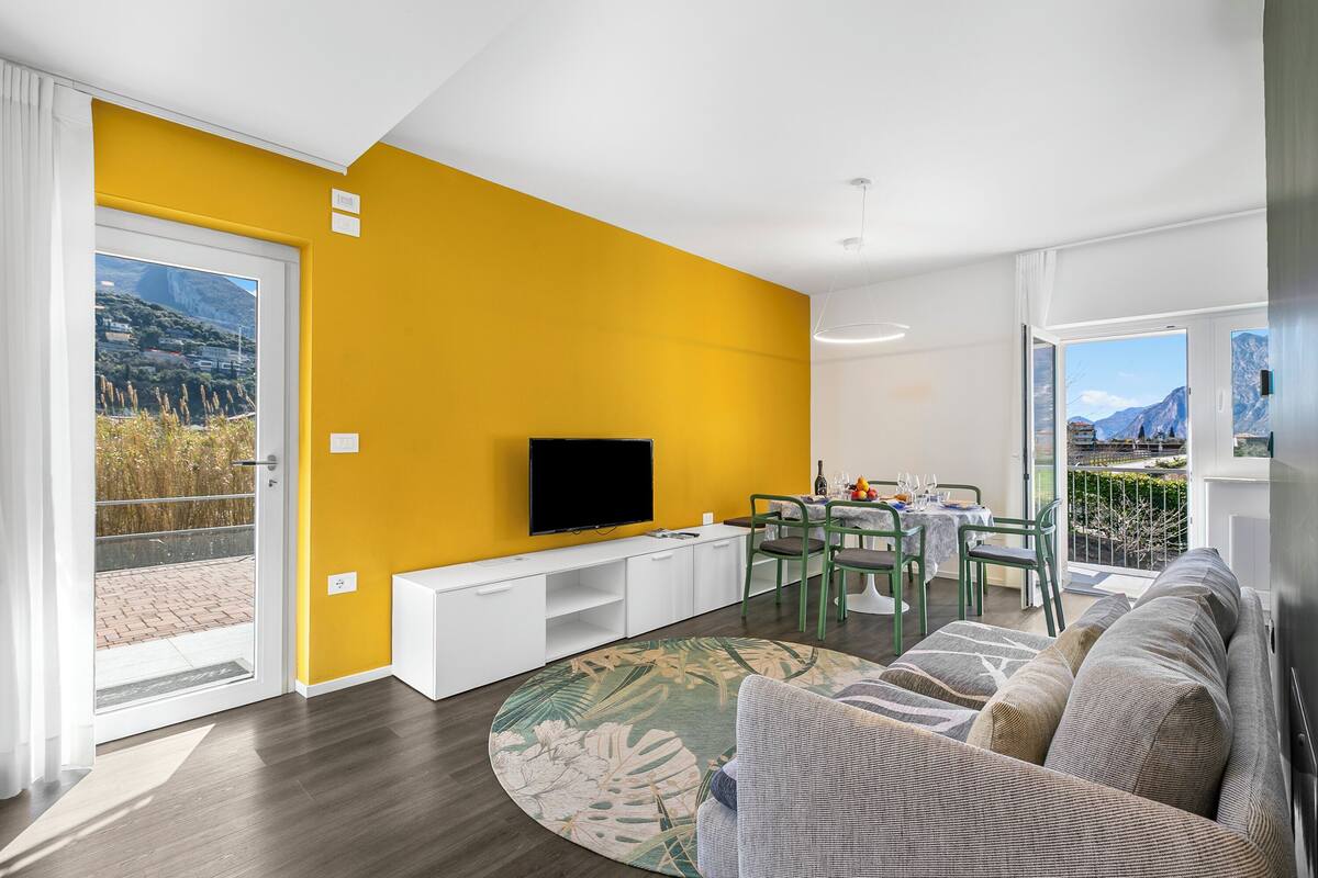 A modern living room with a yellow wall, a flat-screen TV, a sofa, a dining area with a table and chairs, and a view of mountains through the window.