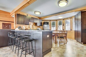 A kitchen with wooden cabinets, a central island with stools, a dining area with a table and chairs, and large windows. - Dock & Views! Lakeside Family Escape in Wilton (Wilton)