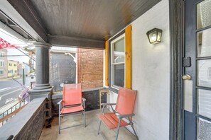 A covered outdoor seating area with two red chairs and a small table, a brick wall, and a window with a yellow frame. - WFH Space & Yard! Walkable Lancaster Row Home (Lancaster)