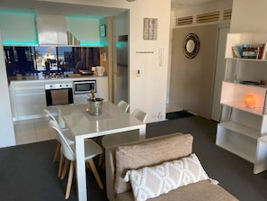 A modern kitchen with white cabinets, a dining table with chairs, and a cozy seating area with a bench and cushions. - Circle on Cavill - Ocean-View Apartment (Surfers Paradise)
