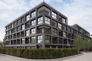 Un edificio moderno a più piani con balconi, ampie finestre e un cespuglio ben curato davanti.