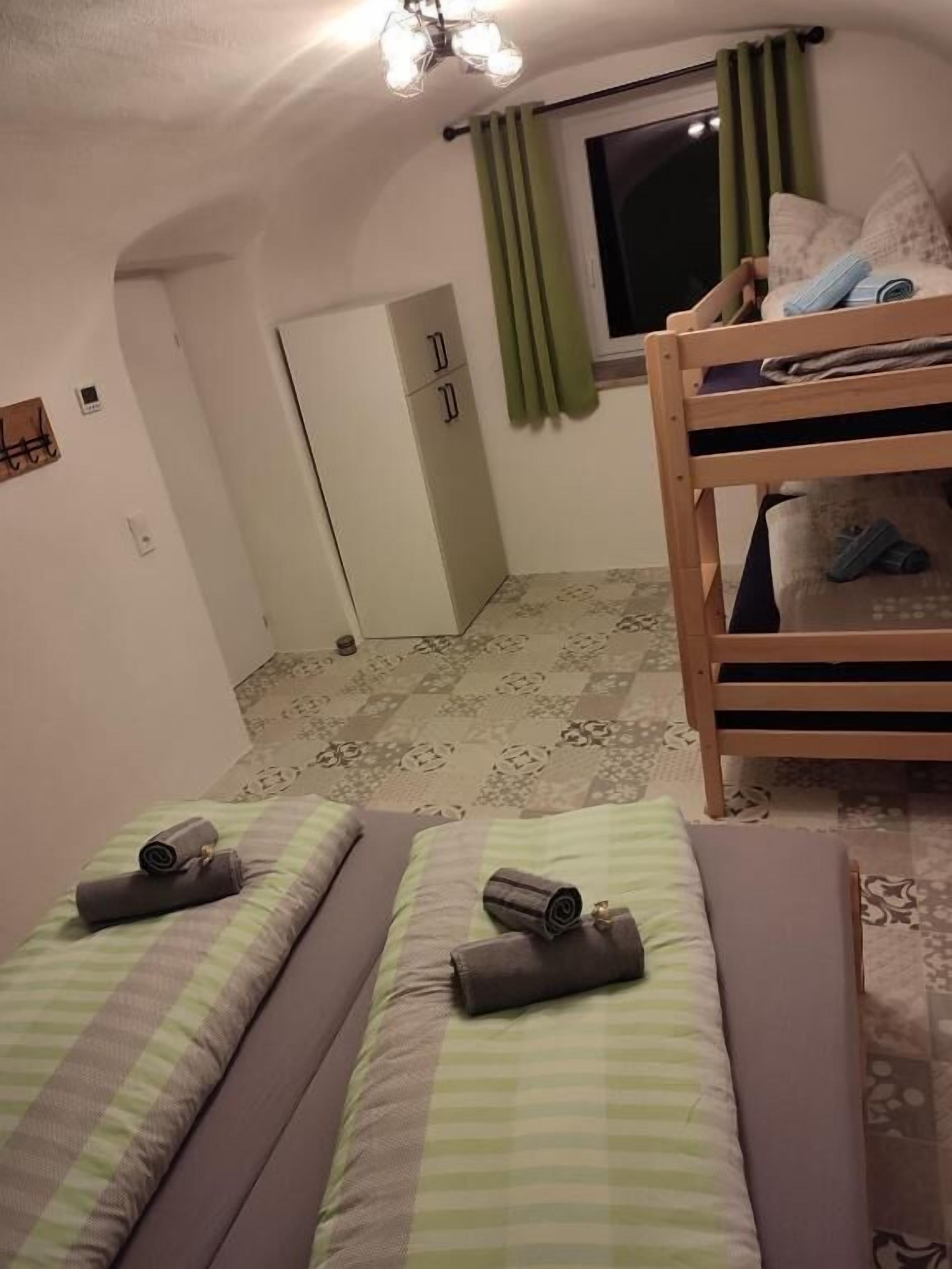 Una stanza piccola e accogliente con due letti singoli, un letto a castello, una finestra con tende verdi e una porta che dà su un'altra stanza.