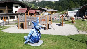Un terrain de jeux avec une statue bleue, des structures de jeu en bois et des bâtiments en arrière-plan.