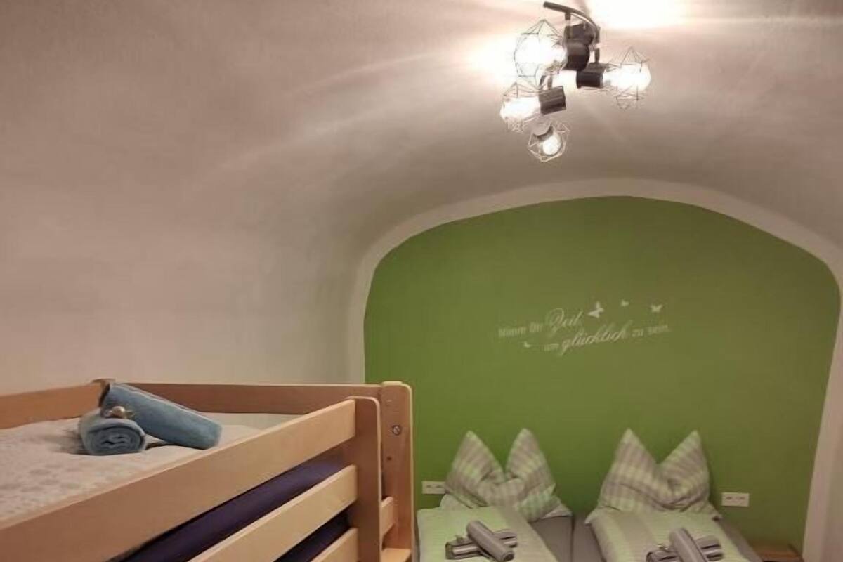 A small, cozy room with a bunk bed and a single bed, both with green bedding. There is a decorative light fixture above the single bed.