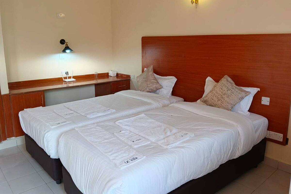 A double bed with white linens and two pillows, a wooden headboard, a bedside table with a lamp, and a wall-mounted light fixture.