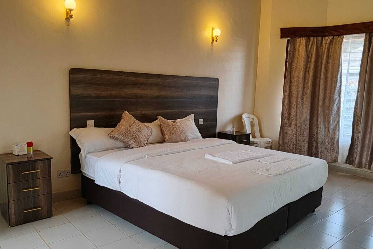 A large bed with a dark headboard, two wall-mounted lights, a nightstand with a bottle, a chair, and a window with curtains.