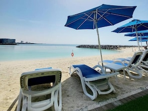 A beach with lounge chairs and blue umbrellas, a clear sky, and a calm sea. - Pacific Sunset Duplex (Jazeerat Al Marjan)