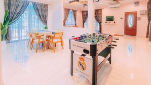 A clean, well-lit room with a foosball table, a dining table with chairs, and large windows with curtains. - Kota Bharu Family Bungalow Homestay (Kota Bharu)