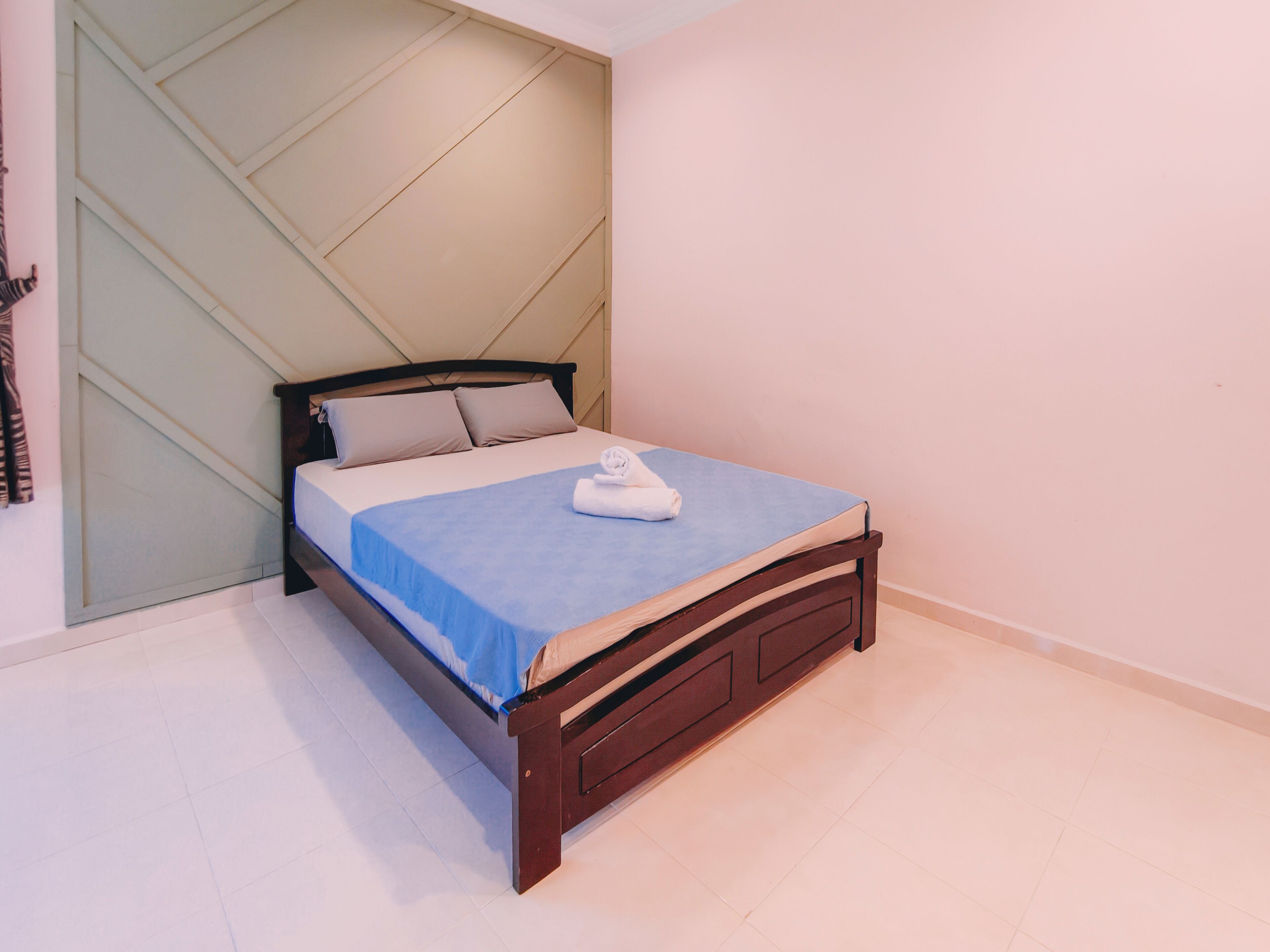 A neatly made bed with a blue bedspread and white pillows, a wooden headboard, and a light-colored wall with a geometric design.