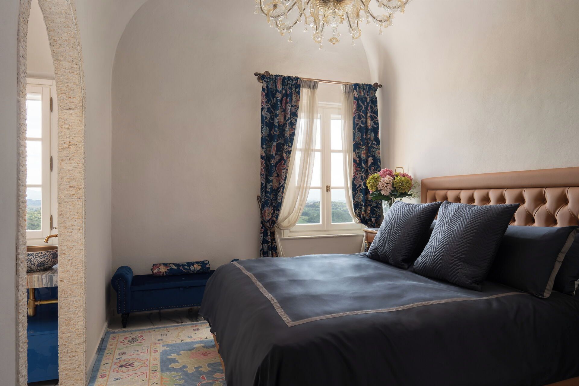 Ein Schlafzimmer mit Bett, einem Fenster mit Vorhängen, einer Kristallleuchtre und Blick ins Freie.