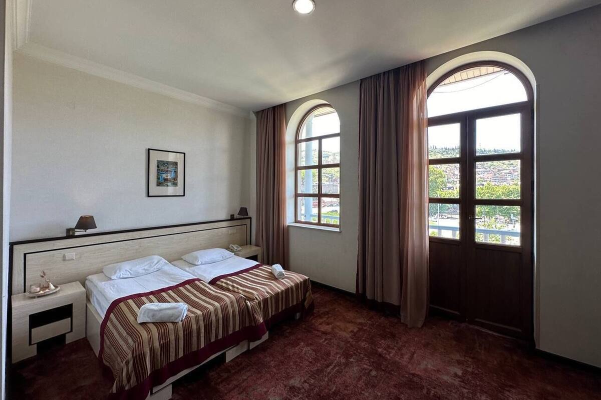 A hotel room with a double bed, a nightstand, a framed picture, and two windows with curtains.