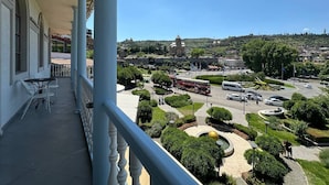 Uma varanda com vista para uma praça, um ônibus e uma colina com uma torre.