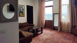 Uma sala de estar com um sofá marrom, uma mesa de centro, um quadro na parede, uma janela grande com cortinas e uma televisão de tela plana.
