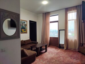 Una sala de estar con un sofá marrón, una mesa de centro, un cuadro en la pared, un ventanal con cortinas y un televisor de pantalla plana.