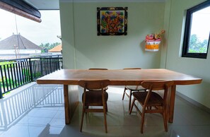 A dining area with a large wooden table and four chairs, a decorative wall hanging, and a window with a view of buildings and greenery. - Toograss Villa Ubud (Ubud)