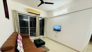 A living room with a brown sofa, a small black table with a plant, a TV mounted on the wall, and a large window with curtains. - 2BHK Service Apart 1703 Parel by Florastays (Mumbai)
