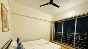 A bedroom with a bed, a ceiling fan, a window with a view of the city at night, and a framed picture on the wall. - 2BHK Service Apart 1703 Parel by Florastays (Mumbai)