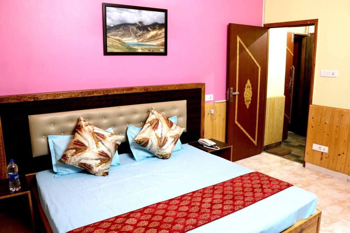 A hotel room with a bed, a nightstand, a red patterned bed runner, a framed picture on the wall, and a wooden door.