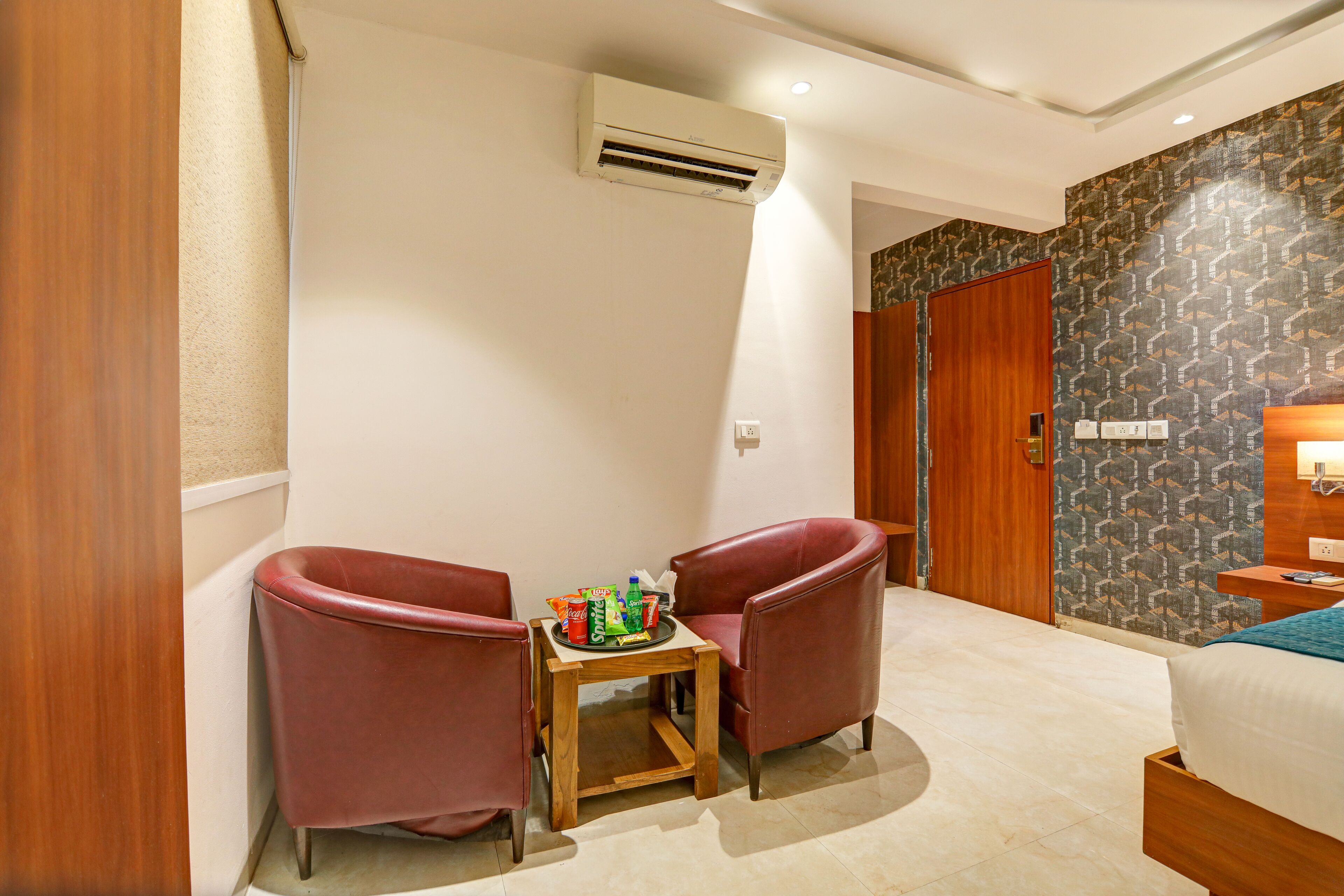 A hotel room with a bed, a wooden nightstand, a table with snacks, two red chairs, and a patterned wall.
