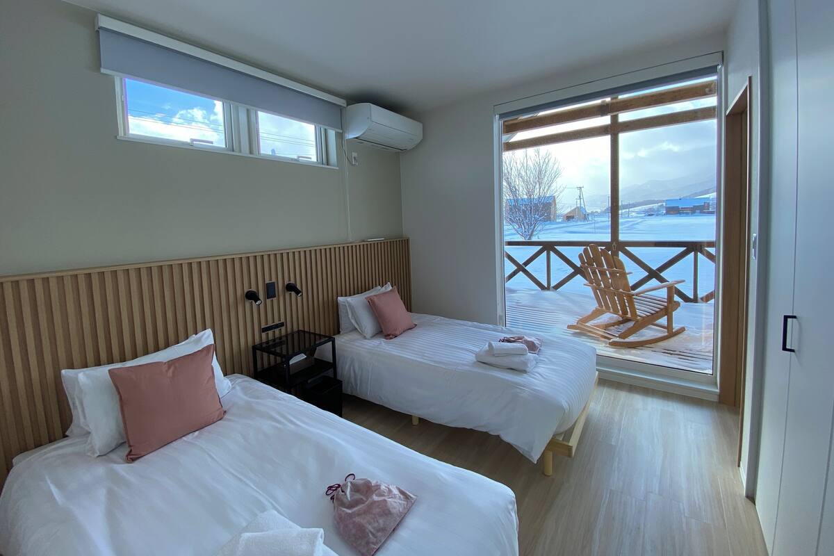Habitación de hotel con dos camas, cabecera de madera, ventana con vistas a un paisaje nevado y balcón con una silla de madera.