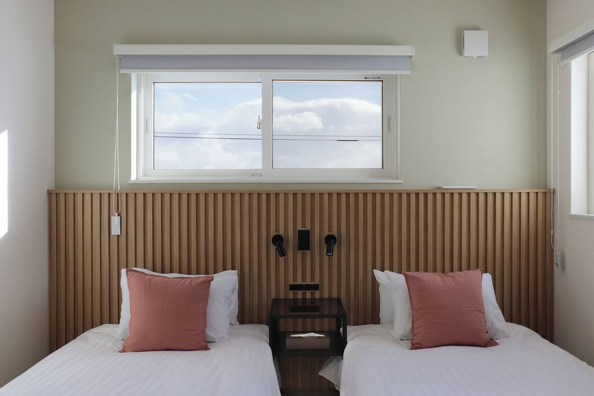 Habitación de hotel con dos camas, una ventana con persianas, cabecero de madera y una mesita de noche con lámpara.