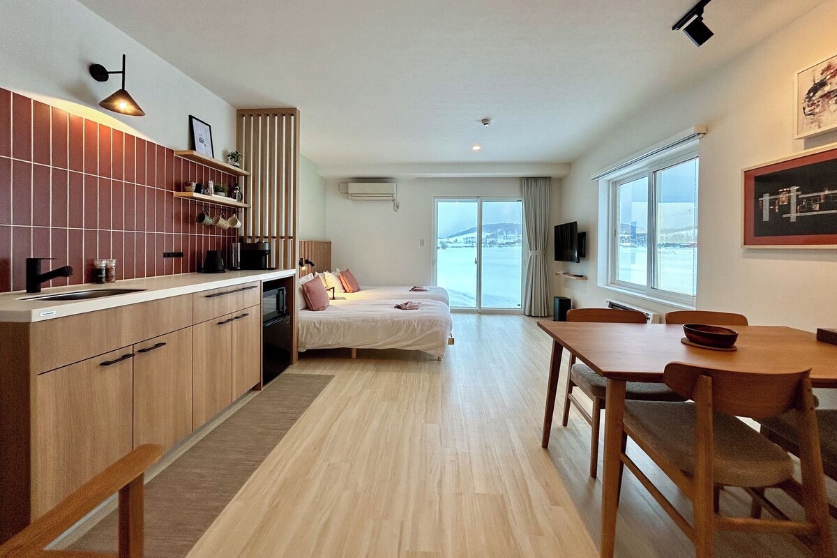 Une cuisine moderne avec des meubles en bois, un coin repas avec une table et des chaises en bois, un lit donnant sur l’extérieur et un téléviseur fixé au mur.