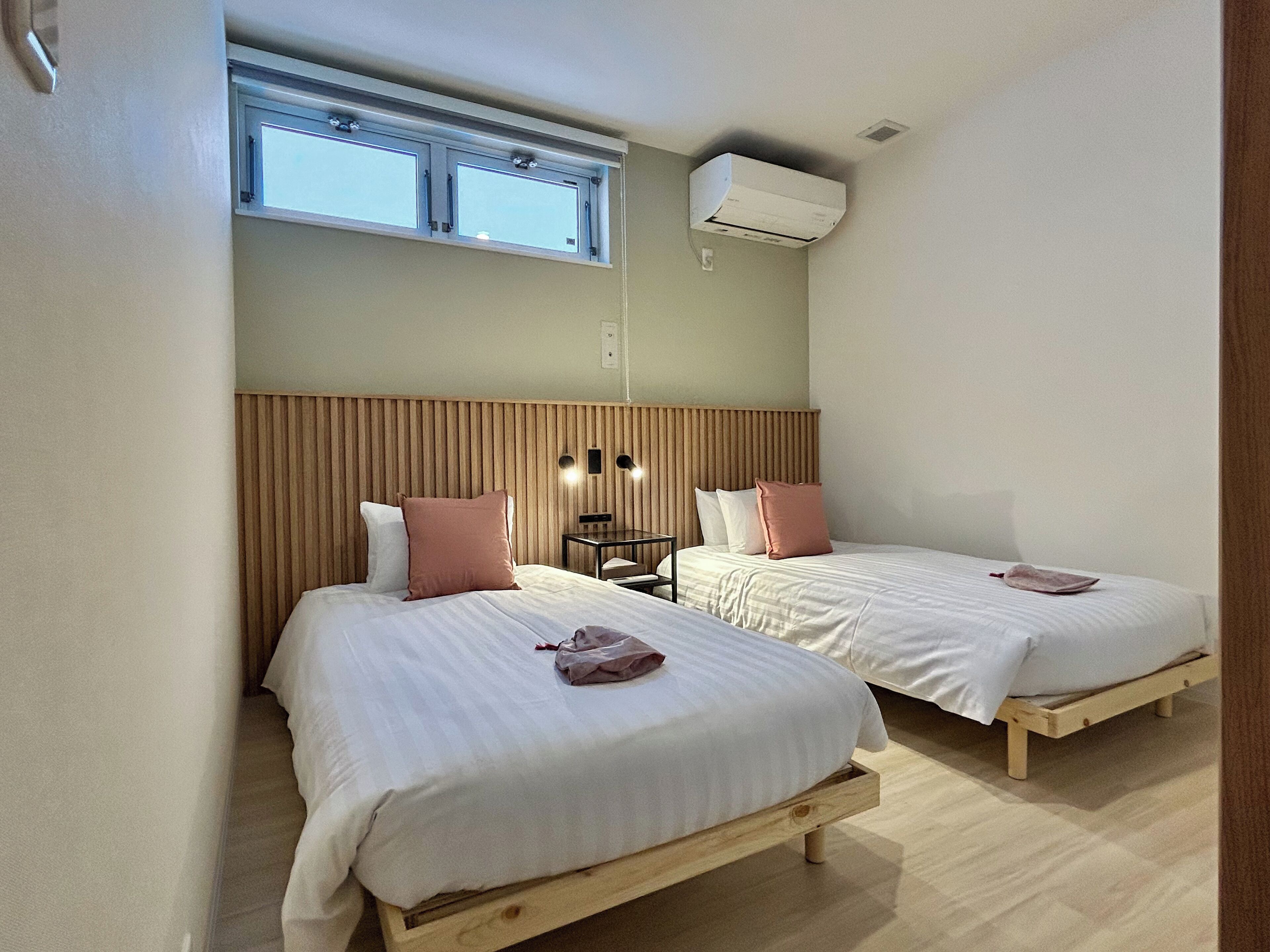 A room with two beds, a wooden headboard, a small table with two lamps, and a window with an air conditioning unit above it.