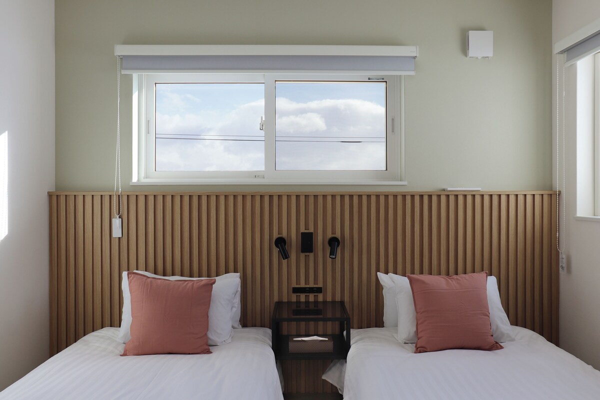 Une chambre d’hôtel avec deux lits, une fenêtre avec des stores, une tête de lit en lattes de bois et une petite table avec une lampe.