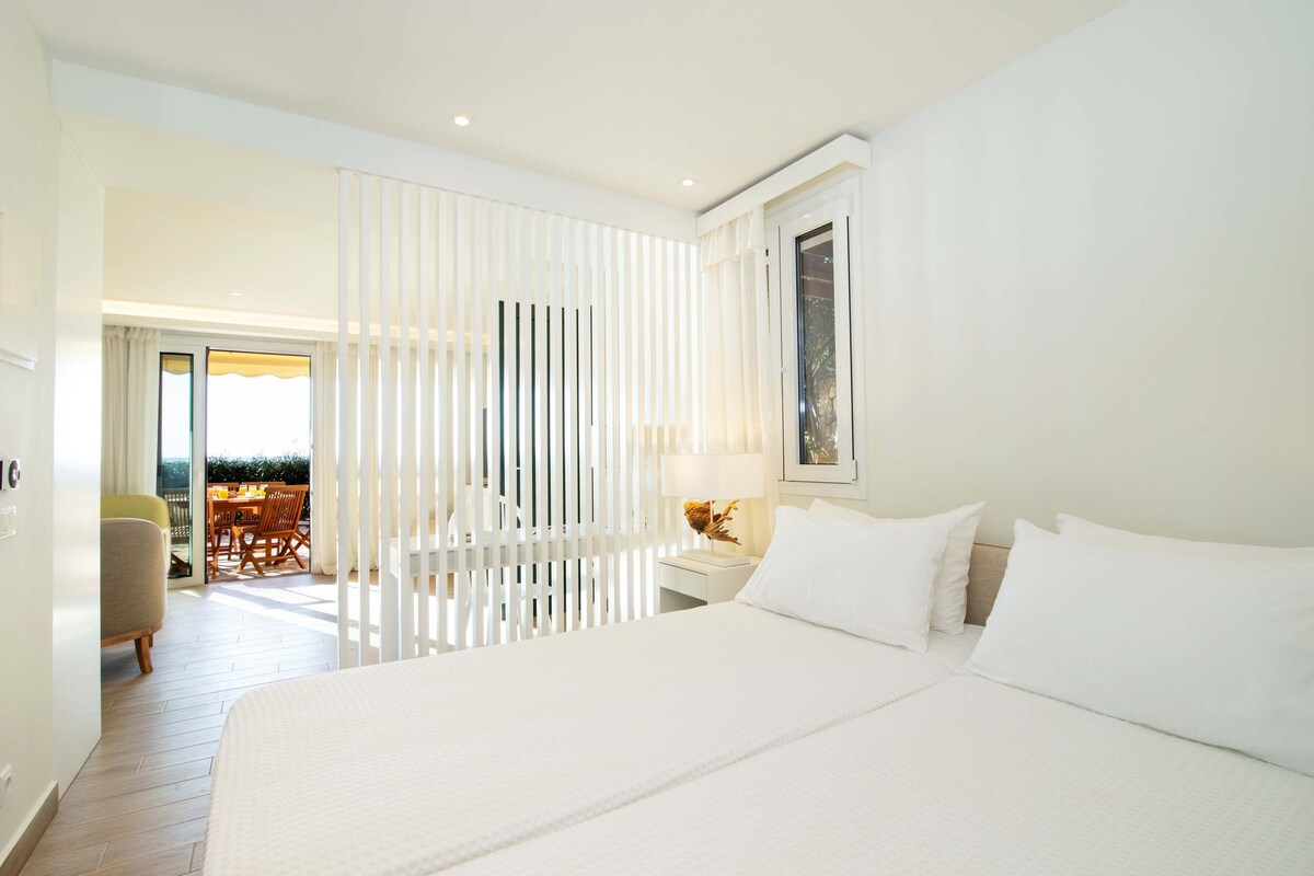 A hotel room with a large bed, a nightstand with a lamp, a chair, and a view of the outdoor terrace.