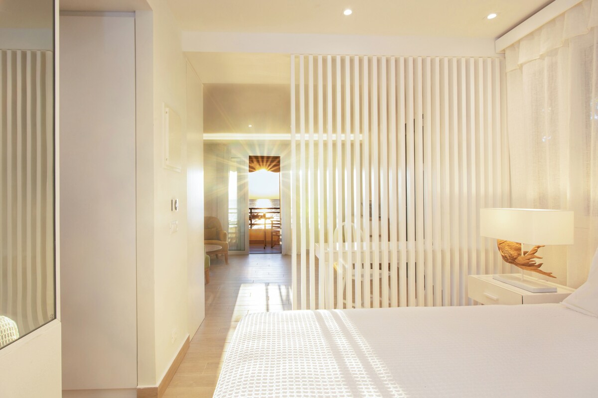 A hotel room with a bed, a nightstand, a lamp, and a view of a hallway leading to a dining area.
