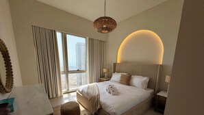 A modern hotel room with a large bed, a round ceiling light, a view of the city through floor-to-ceiling windows, and a small round ottoman. - Top floor Grande 2BR Luxe Panoramic Sea View Bohemian Style (Dubai)