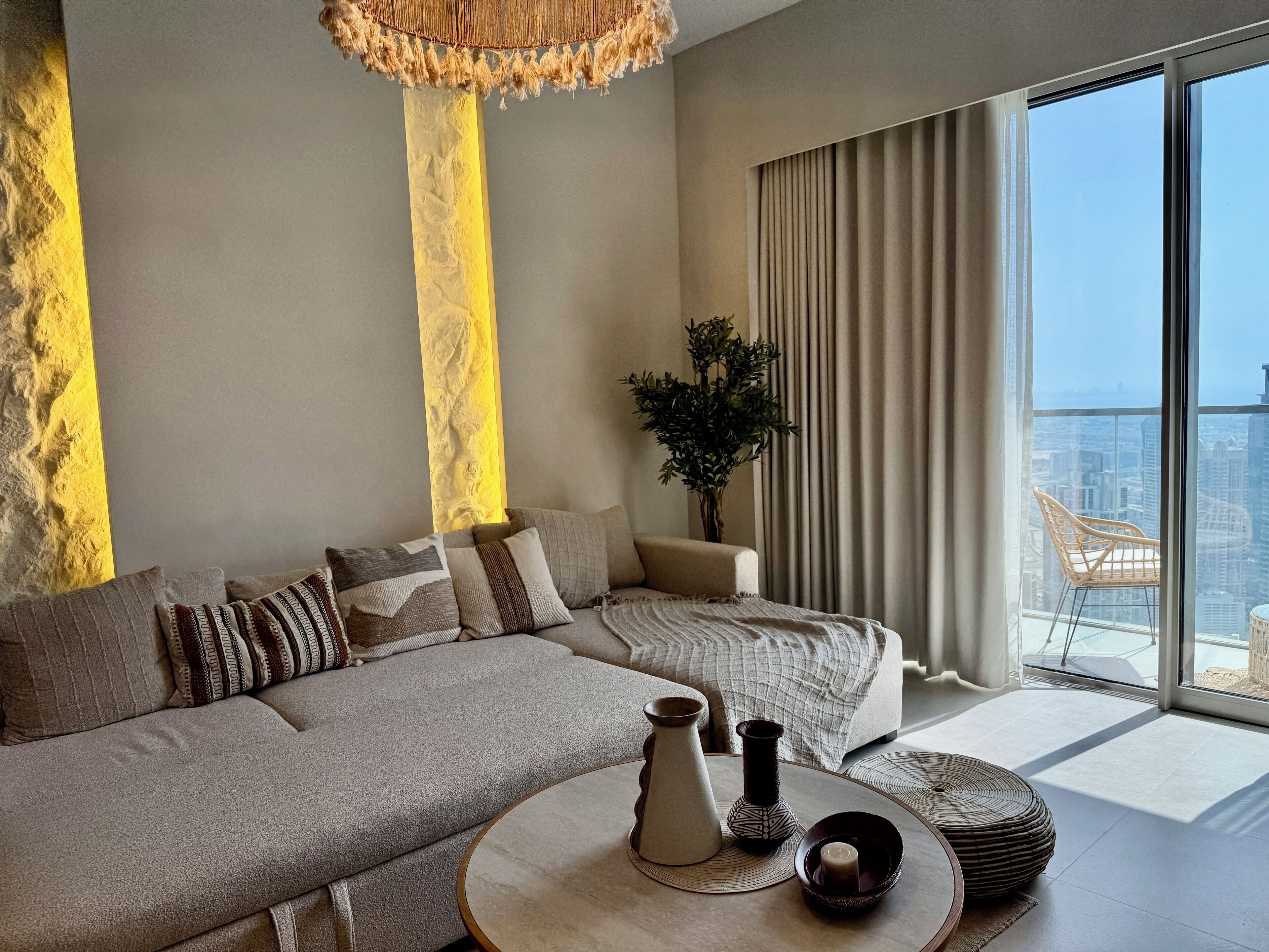 A modern living room with a grey sofa, decorative pillows, a round coffee table with vases, and a view of a cityscape through large windows.