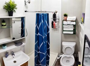 A bathroom with a white sink, a mirror, a shower curtain, a toilet, and a shelf with towels. - Charming Jawn in Fabulous Fishtown (Philadelphia)