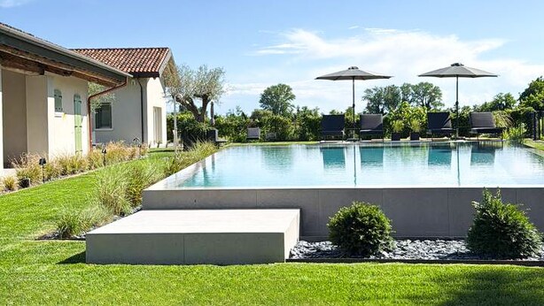 Une piscine avec des chaises longues et des parasols, un bâtiment doté d’un toit en tuiles et un ciel bleu azur.