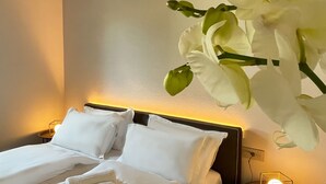 Une chambre d’hôtel avec un lit, des oreillers blancs, une table de chevet avec une lampe et une grande orchidée blanche accrochée au mur.