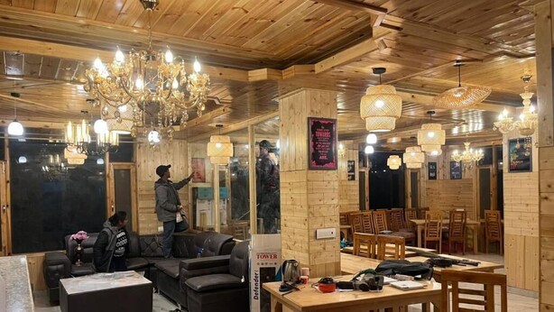 Een gezellige hotellobby met houten wanden en plafond, een kroonluchter, een bank, een zithoek met houten tafels en stoelen, en een bordje voor een evenement met militair thema.