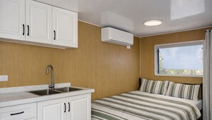A compact room with a bed, a window with curtains, a kitchen area with white cabinets, and an air conditioning unit.