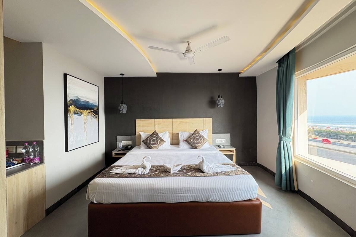 A modern hotel room with a large bed, a ceiling fan, a view of the sea, and a wooden headboard.