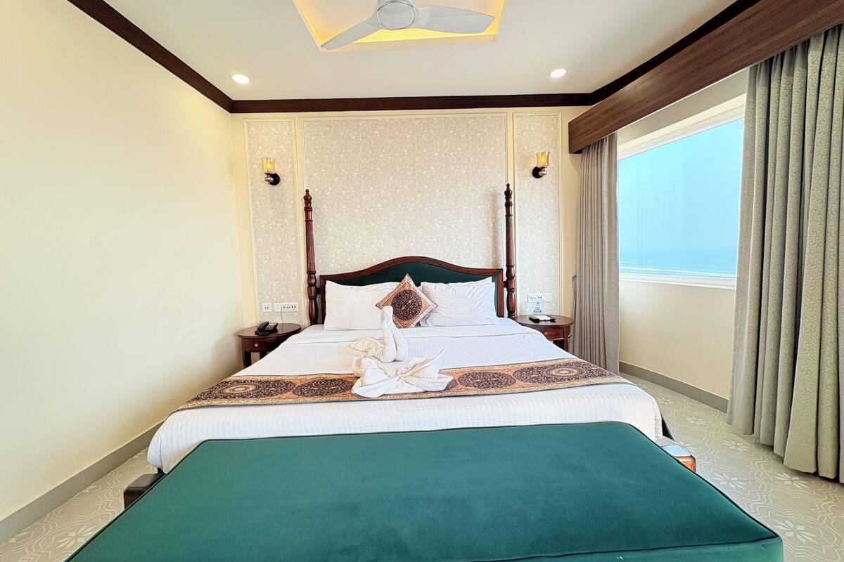 A hotel room with a large bed, a bedside table, a nightstand, a green bench, a ceiling fan, and a window with curtains.